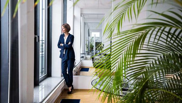 Frau in dunkelblauem Anzug lehnt mit verschränkten Armen am Fenster in einem modernen Büro mit Pflanzen